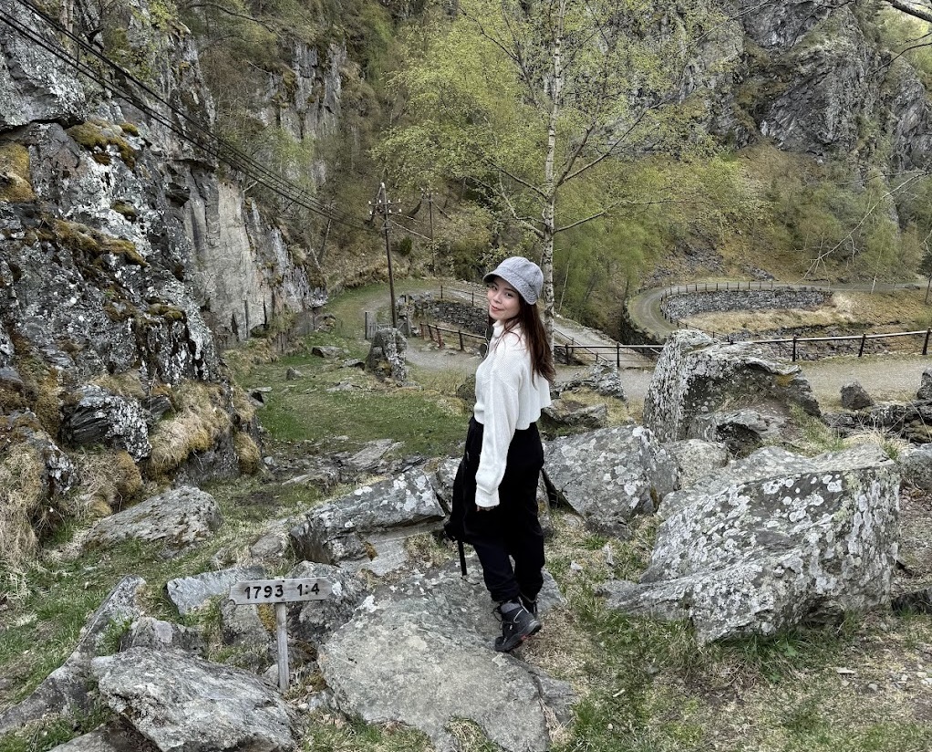 Woman standing on rocks near a historic 1793 marker along the winding Vindhellavegen trail in Lærdal, Norway, with stone curves and cliffs in the background.